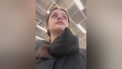 Media: Video of a young woman with straight brown hair, wearing a black puffer jacket, looking up in an indoor setting with white ceiling tiles and fluorescent lighting.