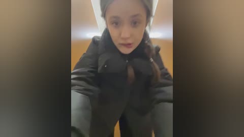 Media: A video of a young Asian woman with light skin and brown hair, wearing a black puffer jacket, standing in a dimly lit room.