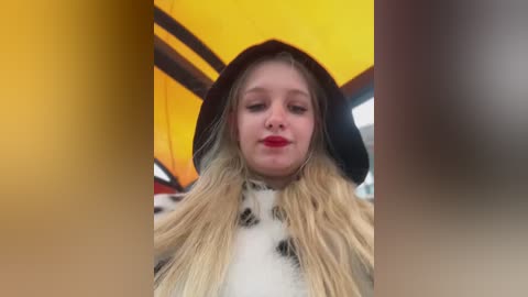 A video of a young woman with long blonde hair, wearing a black hooded jacket and a white and black polka dot dress, under a yellow and black striped umbrella, with a blurred background.