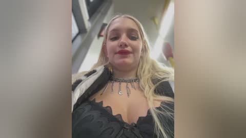 Media: A video of a blonde woman with fair skin, wearing a black lace bra and silver choker necklace, smiling confidently. The background is a blurry subway car with yellow caution tape.