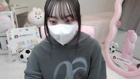 Media: Video of an Asian girl with straight black hair and fair skin, wearing a gray hoodie with a \"2\" logo and a white face mask, sitting in a pastel-themed room filled with plush toys and a pink mirror.