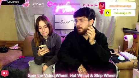 Video of two young people sitting on a couch, watching a video on their phones. Background features a pink neon sign and messy room with a keyboard. Text overlay: \"Live from the Video Whore, Hot Whirl & Strip Wheel!\