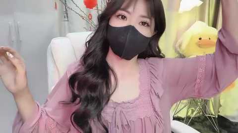 Media: Video of an Asian woman with long black hair, wearing a black face mask and lavender blouse, sitting in a modern room with yellow curtains and a yellow duck toy.