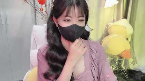 Media: Video of an East Asian woman with long, wavy black hair, wearing a black face mask, a lavender blouse, and a yellow stuffed toy. Background includes a white chair, a yellow lamp, and a floral wall painting.