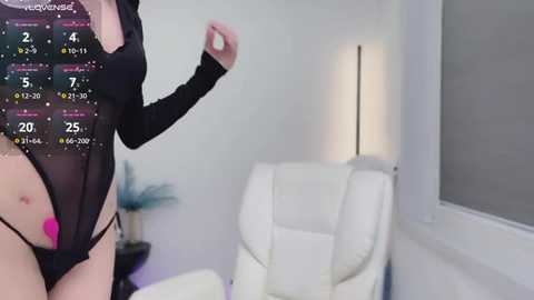 Media: Video of a woman in a sheer black bodysuit, standing in a minimalist white room with a modern lamp and white chair.