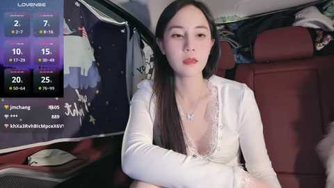 Media: A video of an East Asian woman with long black hair, fair skin, and red lipstick, wearing a white lace-up top, sitting in a red leather car seat, with a digital display showing weather and temperature information.