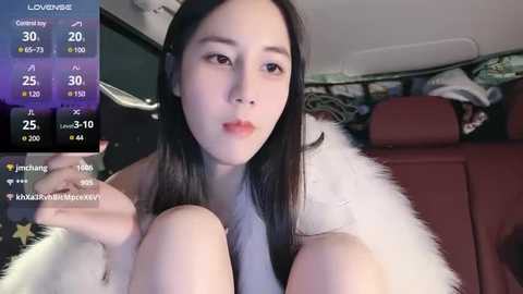 A video of an East Asian woman with straight black hair, light skin, and full lips, sitting in a car, wearing a white fur coat, with a video overlay displaying live streaming statistics.