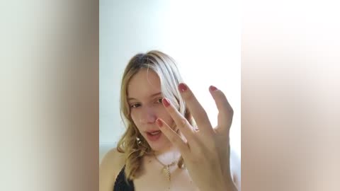 Media: Video of a blonde woman with fair skin and red nails, wearing a black top, standing against a bright white background, her left hand raised.