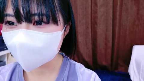 Media: Video of an East Asian woman with short black hair, wearing a white surgical mask, light blue scrubs, and a white headband. Background features brown curtains and a white medical table.