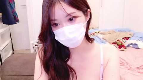 Media: Video of an East Asian woman with long, wavy brown hair, wearing a white face mask, light pink bra, and sitting on a bed with clothes scattered around.