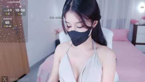 Media: A video of an Asian woman with long black hair, wearing a black face mask, a white halter top, and a pink blanket in a modern, lightly furnished bedroom.