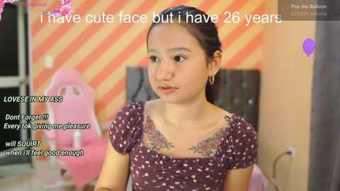 Media: Video of a young Asian woman with fair skin, dark hair, and a floral tattoo on her chest, wearing a brown dress. Text overlays her face, saying \"I have cute face but I have 26 years.\" Background shows a pink gaming chair and orange striped wall.