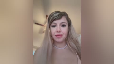 Media: Video of a young woman with light skin and long, straight blonde hair, wearing a white top and pearl necklace. She has a soft smile and sits in a dimly lit room with beige walls and a ceiling fan.