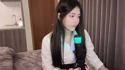 Media: Video of an East Asian woman with long black hair, wearing a white shirt, black shoulder harness, and a green oxygen mask, sitting on a beige bed in a hotel room with a brown curtain and a kitchenette in the background.