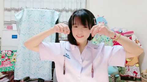 Media: Video of an Asian woman with shoulder-length black hair, wearing a white button-down shirt, smiling and playfully pulling her earlobes in a cluttered, colorful bedroom.