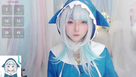 Media: Video of a fair-skinned, young woman in a blue and white shark cosplay, with long, pastel-colored hair, standing in a softly lit room with a calendar overlay.