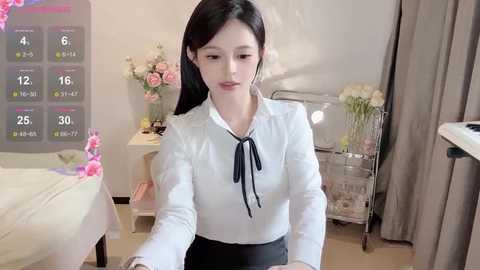 Media: Video of a slender East Asian woman with long black hair, wearing a white blouse and black skirt, sitting on a bed in a pastel-toned room with flowers, calendar, and a metal cart.