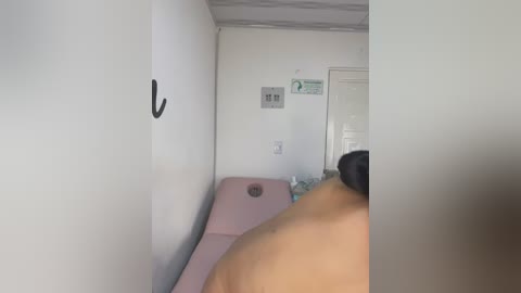 Media: Video of a narrow, white-walled room with a pink examination table. A person with a tattoo on their shoulder is partially visible, and there's a black hook on the wall.