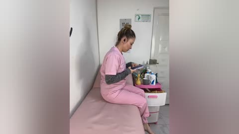 Media: Video of a light-skinned woman with blonde hair in a bun, wearing pink scrubs, sitting on a pink bed, reading a tablet, in a sparsely furnished, white-walled hospital room with a closet and a plastic laundry basket.