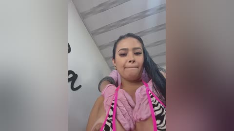 Media: Video of a Latina woman with medium-dark skin and long dark hair in a pink and black zebra-patterned top, holding a pink towel. She stands in a white-walled hallway with black graffiti.