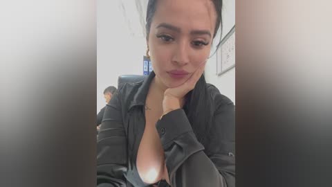 Media: Video of a Latina woman with medium skin tone, dark hair, and full lips, wearing a black leather jacket, seated at a desk with a blurred background.