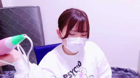 Media: Video of an Asian woman with short brown hair wearing a white mask, holding a pink and green hairdryer, seated on a grey, quilted chair with a blue cushion.