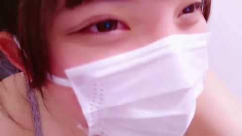 Media: Close-up video of an East Asian woman with fair skin, wearing a white surgical mask partially covering her nose and mouth, revealing light brown eyes and straight black hair.