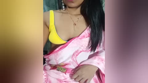 Media: Video of a woman with medium brown skin, wearing a yellow bra and pink sari with floral embroidery, adorned with a gold necklace. The background is blurred, featuring a gradient of yellow, purple, and pink.