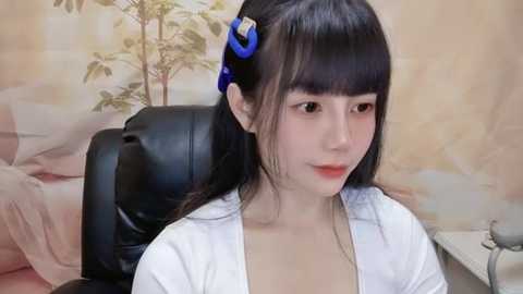 Media: A video of an East Asian woman with straight black hair, styled with a blue hair tie, wearing a white blouse, sitting in a black leather office chair.