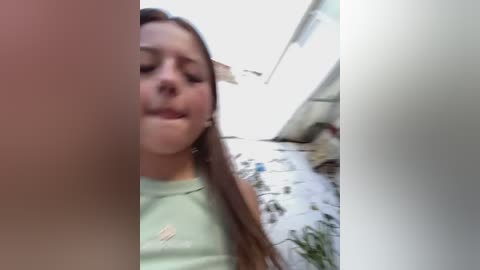 Media: Video of a young woman with long brown hair and a light green top, looking downward with a neutral expression, taken in a small, dimly lit room with a white ceiling and a plant in the background.