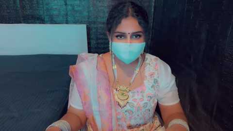 Media: Video of a South Asian woman in traditional saree with floral patterns, wearing a blue face mask, sitting on a dark couch against a brick wall.