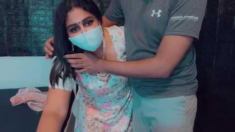 Media: Video of a South Asian woman with a mask, wearing a floral dress, being held by a man in a gray shirt, in a dimly lit room with a brick wall.