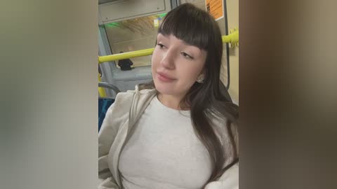 Media: Video of a young Asian woman with long black hair and a light beige jacket, sitting on a yellow subway seat, smiling slightly. Background shows subway interior with windows and yellow railings.