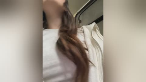 Media: A close-up video of a person with long, wavy brown hair, wearing a white shirt. The image is slightly blurry and the background is indistinct.