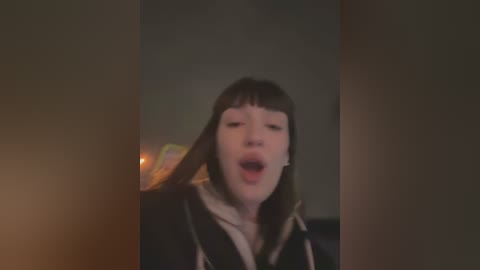 Media: A blurry, low-light video of a young person with straight, shoulder-length dark hair, wearing a dark hoodie, singing passionately with eyes closed, against a dimly lit background.