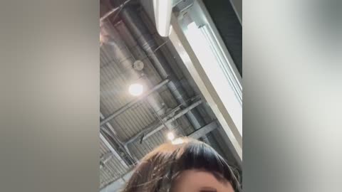 Media: A low-angle video captures a woman with short, dark hair, her face partially obscured, standing in an industrial room with exposed pipes, fluorescent lighting, and a metal ceiling.