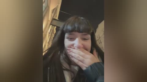 Media: A blurry video of a young woman with long dark hair, wearing a black jacket, covering her mouth with one hand. The background shows a dimly lit room with beige walls.