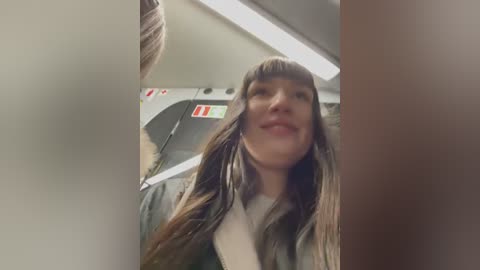 Media: Video of a young woman with long brown hair, wearing a green jacket, smiling inside a train compartment with fluorescent lighting, blurred background, and an out-of-focus person visible.