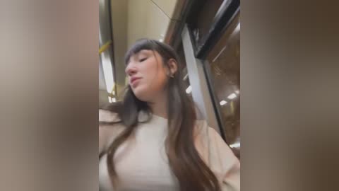 Media: A video of a young woman with long, straight brown hair, wearing a beige top, leaning against a glass elevator. The background features modern interior design with metallic accents.