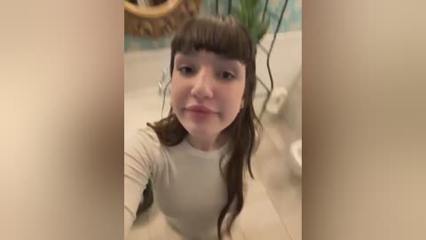 Media: A video of a young woman with long brown hair and bangs, wearing a white long-sleeve shirt, standing in a bathroom with a wicker basket and green plants in the background.