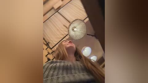 Media: Video of a young woman with long blonde hair, wearing a gray sweater, standing in a dimly lit room with wooden walls and a circular light fixture.