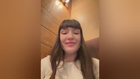 Media: Video of a young woman with long, straight brown hair and bangs, wearing a white top, smiling. She is in a wooden room with warm lighting and a wooden ceiling.