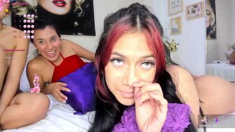 Media: Video of a young woman with long black hair, red highlights, and light skin, lying on a bed with a purple pillow, smiling seductively. Two other women, one in a red top, the other topless, are in the background.