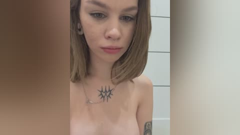 Video of a topless young woman with light skin and straight, shoulder-length brown hair. She has a tattoo of a black starburst on her chest and a tattoo on her left arm. The background features white tiles and a blurred figure.