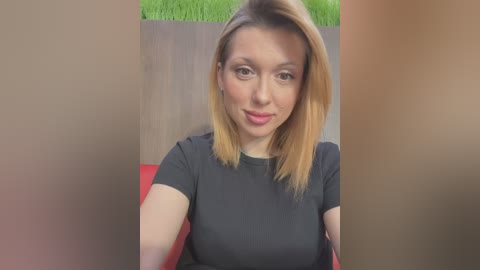 Media: Video of a young Caucasian woman with straight, shoulder-length blonde hair, wearing a black t-shirt, seated indoors against a wooden backdrop.