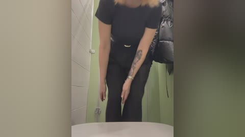 Media: Video of a woman in a bathroom, wearing black pants and a black top, with a black jacket hanging on the wall, and a tattoo on her left arm.