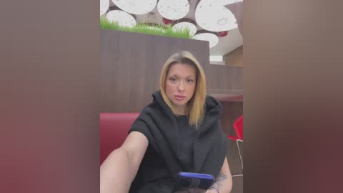 Video of a blonde woman with light skin, wearing a black hoodie, seated in a modern, red-cushioned booth. She holds a blue smartphone. Background features a beige wall with white, circular light fixtures and greenery.