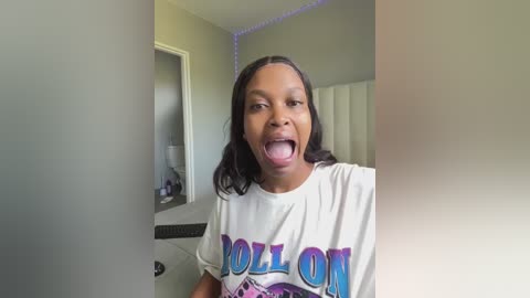 Media: A video of a young Black woman with medium-brown skin and long, straight black hair, wearing a white \"Beyonc\u00e9\" t-shirt, sticking her tongue out in a playful manner. The room has a green wall and a white cabinet with purple lights in the background.