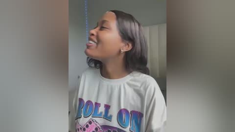 Media: Video of a young woman with medium brown skin, shoulder-length dark hair, wearing a white t-shirt with \"POLL ON\" in blue and purple text, laughing while leaning against a wall.