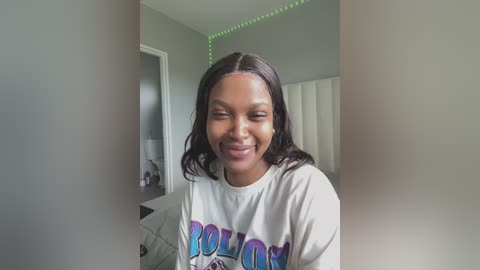 Media: Video of a smiling Black woman with medium skin tone and shoulder-length black hair, wearing a white T-shirt with colorful \"Polo\" text, in a dimly lit, gray-walled room with a green LED strip light on the ceiling.